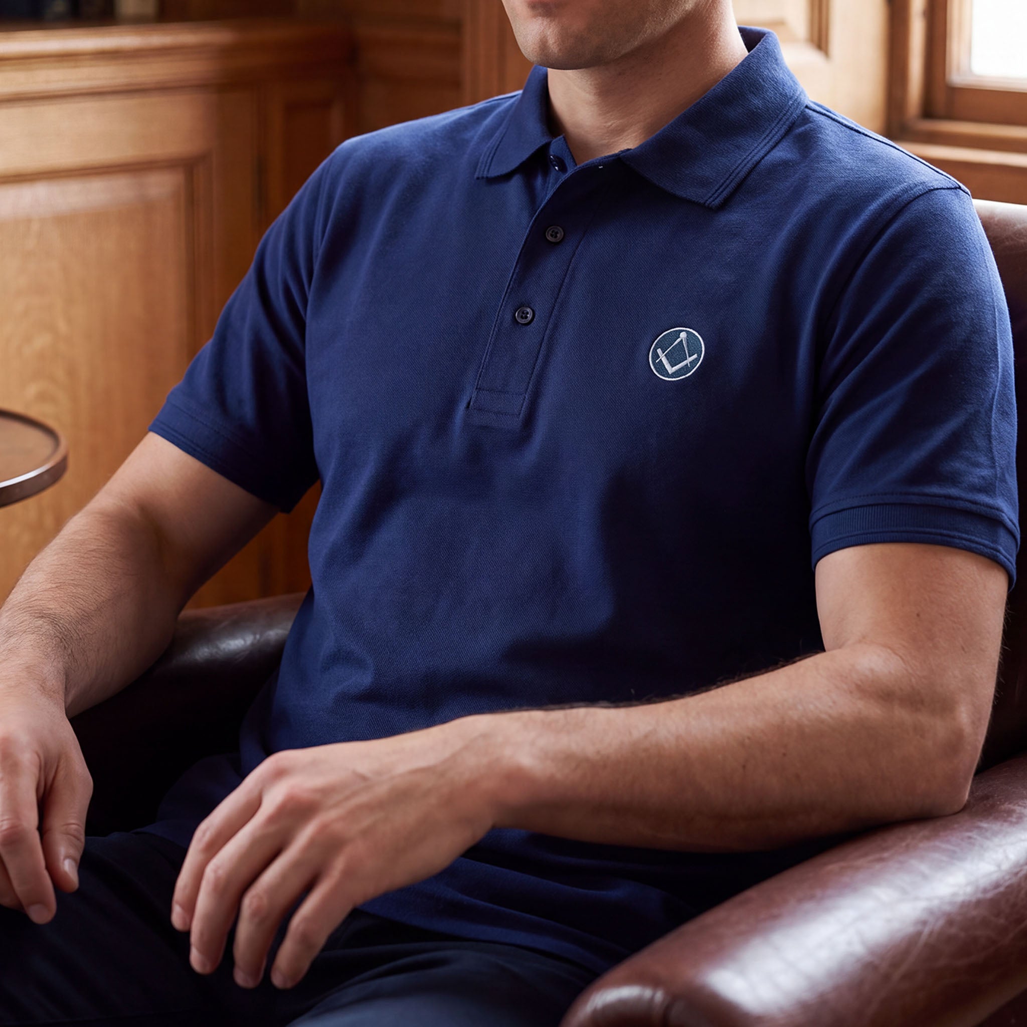 Model wearing blue polo shirt with a collar and button placket, featuring a small sliver and blue embroidered Masonic square-and-compass symbol on the chest.