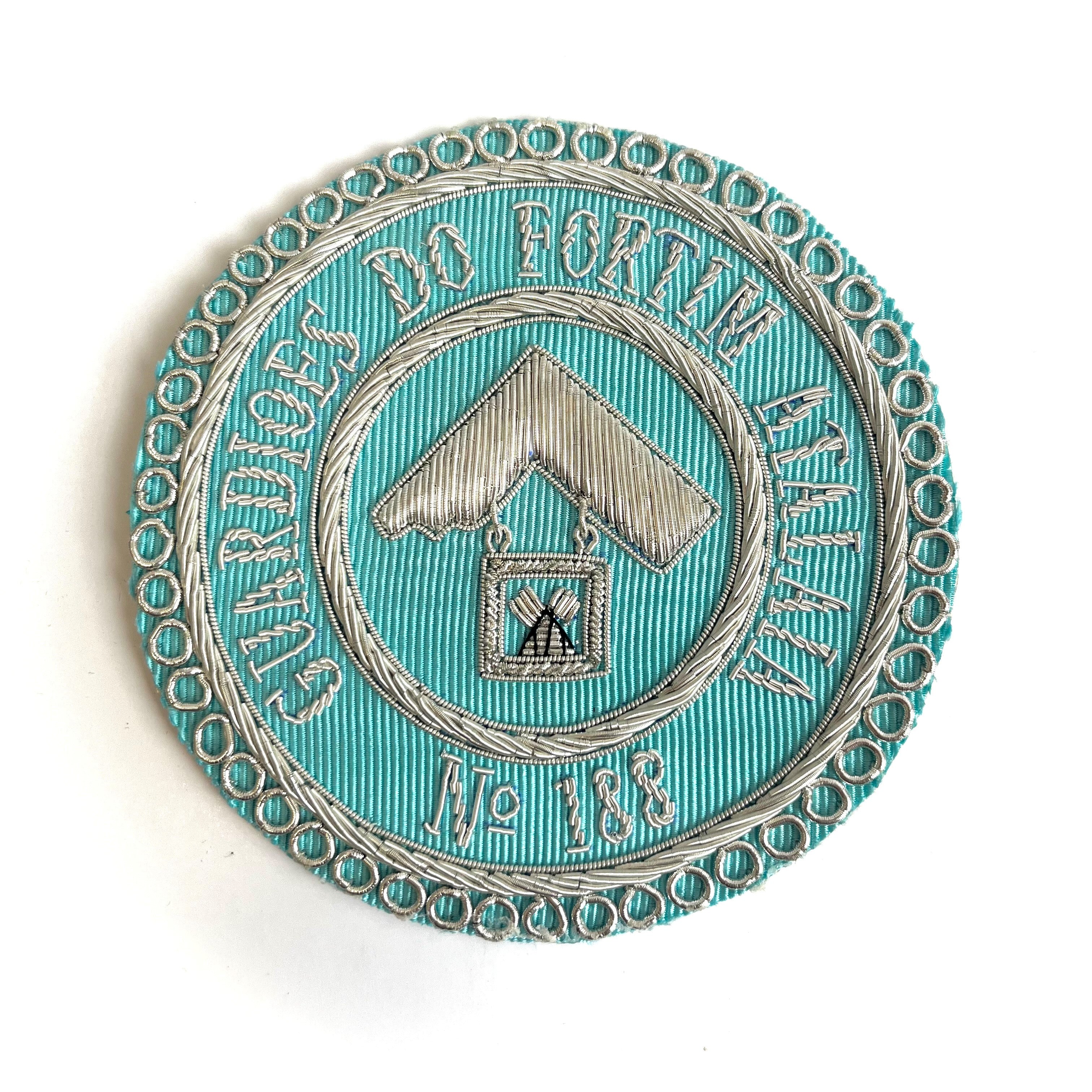 A Craft Light Blue Masonic Apron Badges circular with embroidered text and a symbol in the center, with no visible apron attachment.
