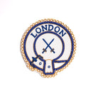 A gold-colored Mark District Undress Apron Badge with the word 'LONDON' and an emblem embroidered on it, without adhesive backing.