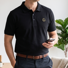 Model wearing black polo shirt with a collar and button placket, featuring a small gold embroidered Masonic square-and-compass symbol on the chest.