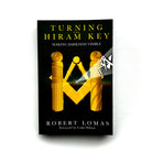 This image features the cover of the book titled "Turning the Hiram Key" by Robert Lomas. The cover design is elegant and mysterious, displaying a dark, foreboding background with a prominent, golden Masonic emblem and a pair of hands holding a light above it.